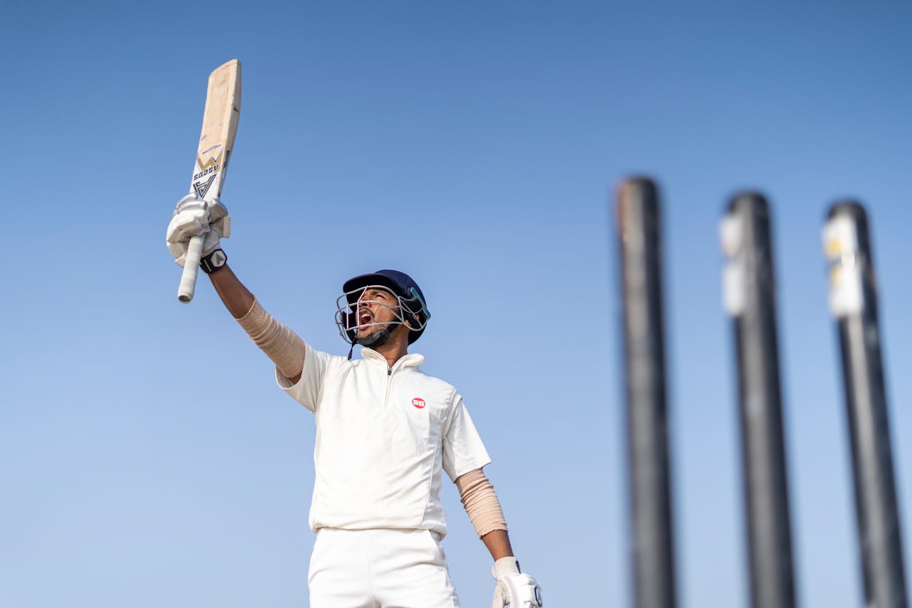 gallery-6 A cricket player exults in victory, raising his bat skyward amidst clear skies.