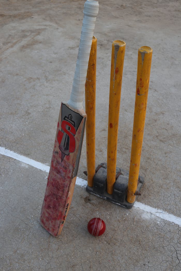 services-02 Still life of a cricket bat, red ball, and stumps on a cricket field, highlighting the sport's essential equipment.