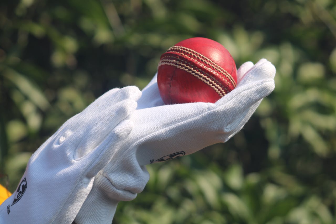hero-img-01 Detailed photo of a red cricket ball held by gloved hands outdoors.